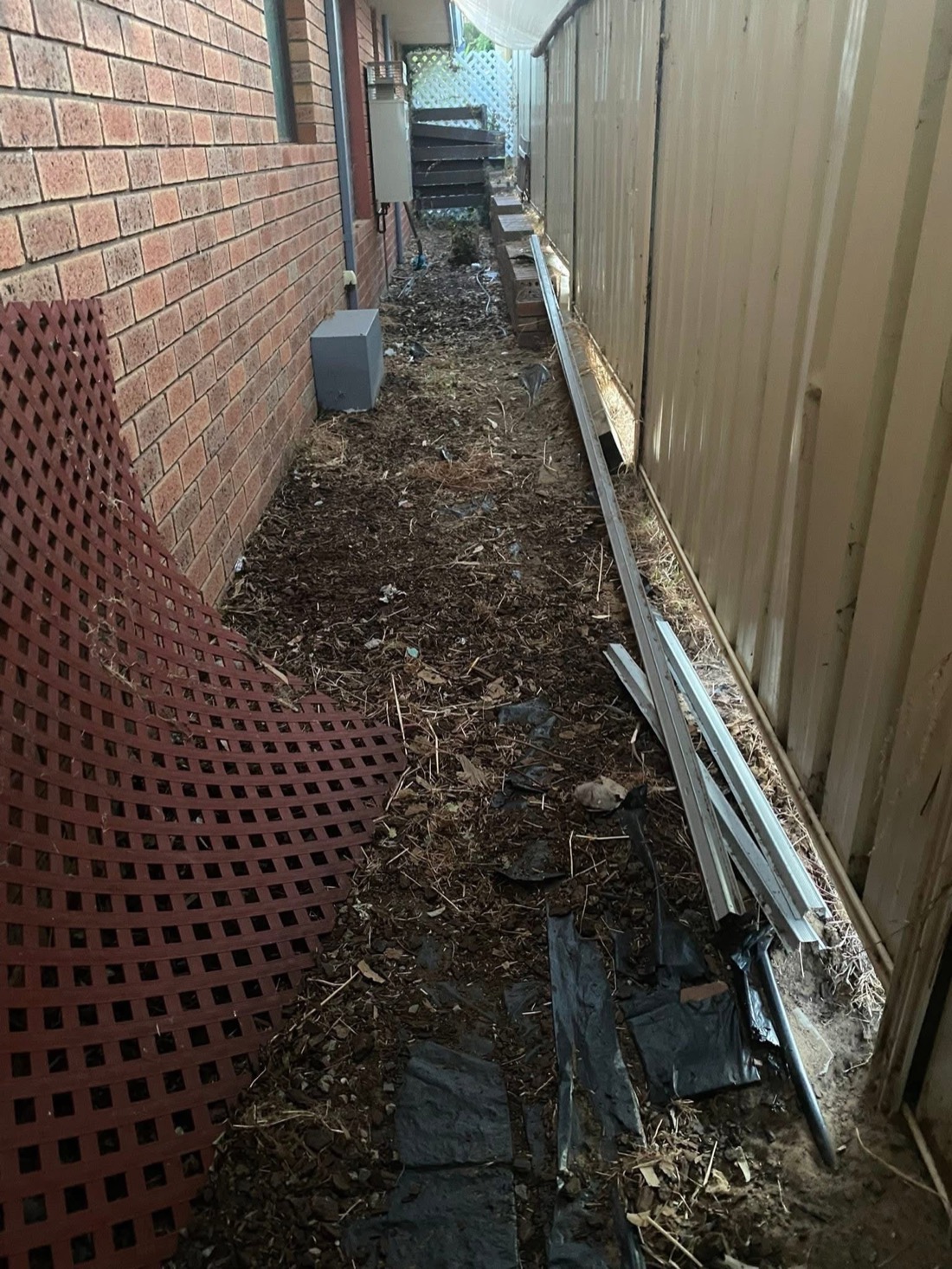 A messy side passage with dirt, debris and discarded materials between a brick wall and a metal fence, before installation.