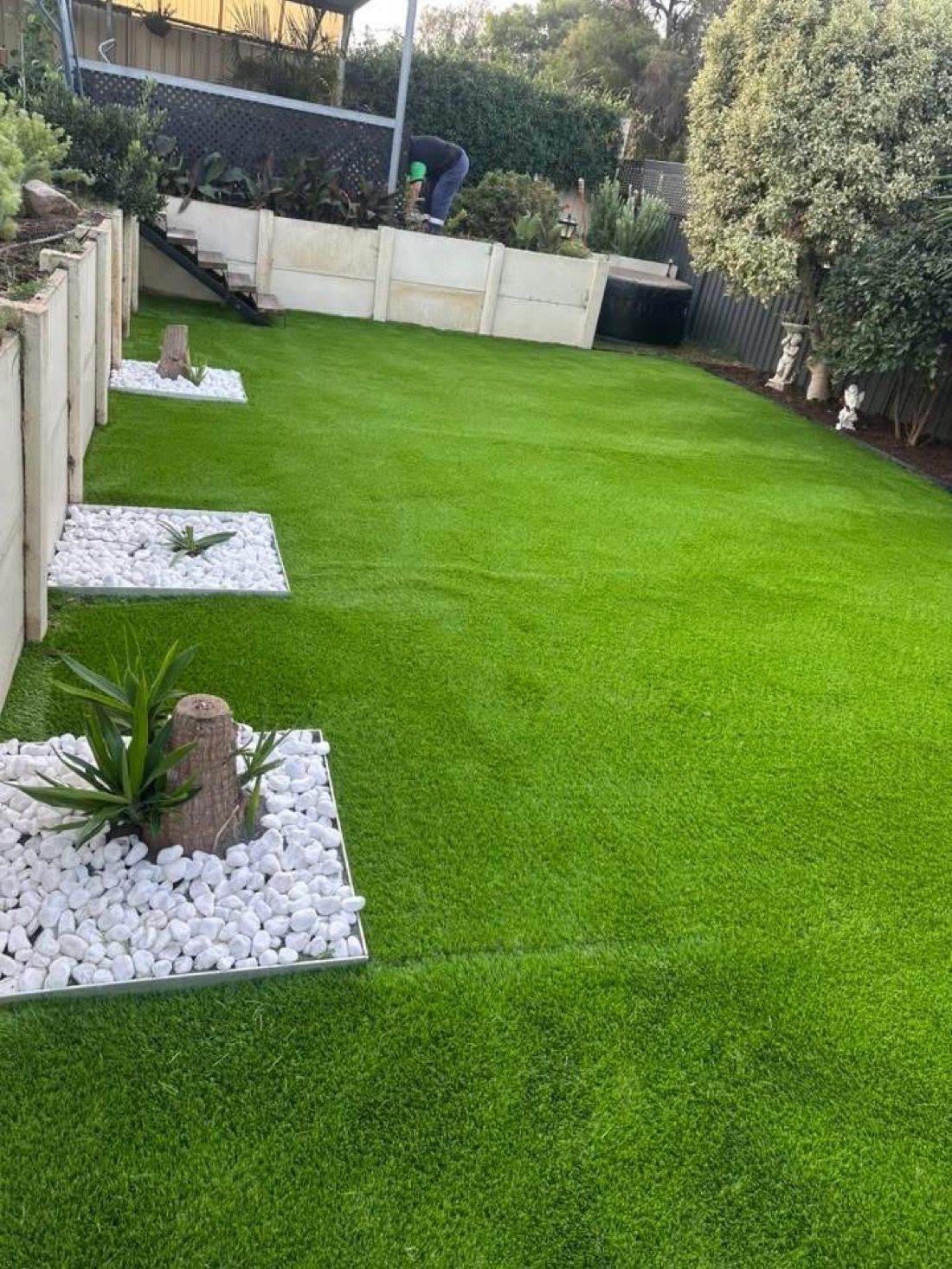 The same backyard transformed with lush green turf, stepping-stone garden islands and white pebble borders, after installation.